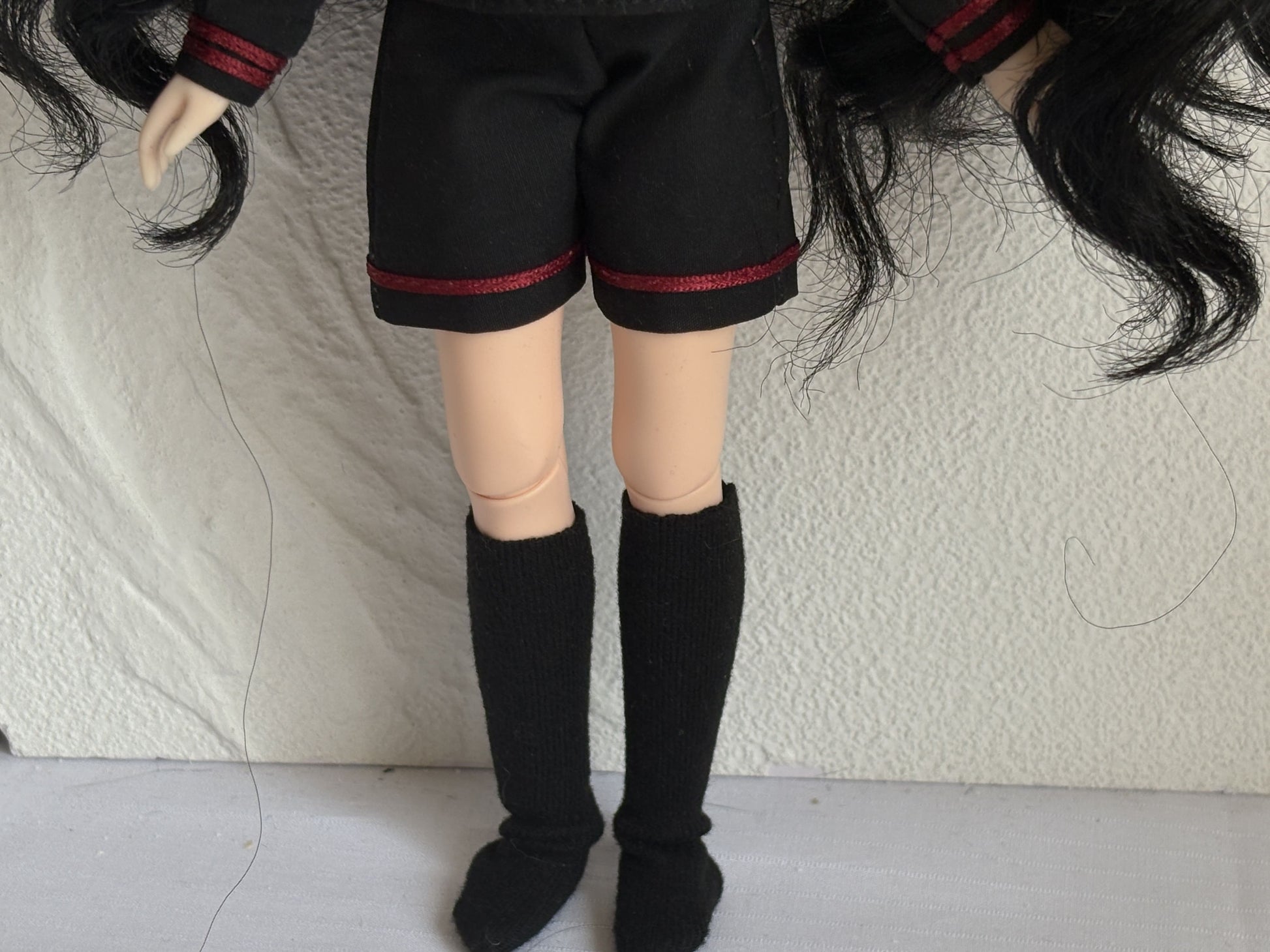 Doll wearing black shorts with red trim and black knee-high boots on a textured white background