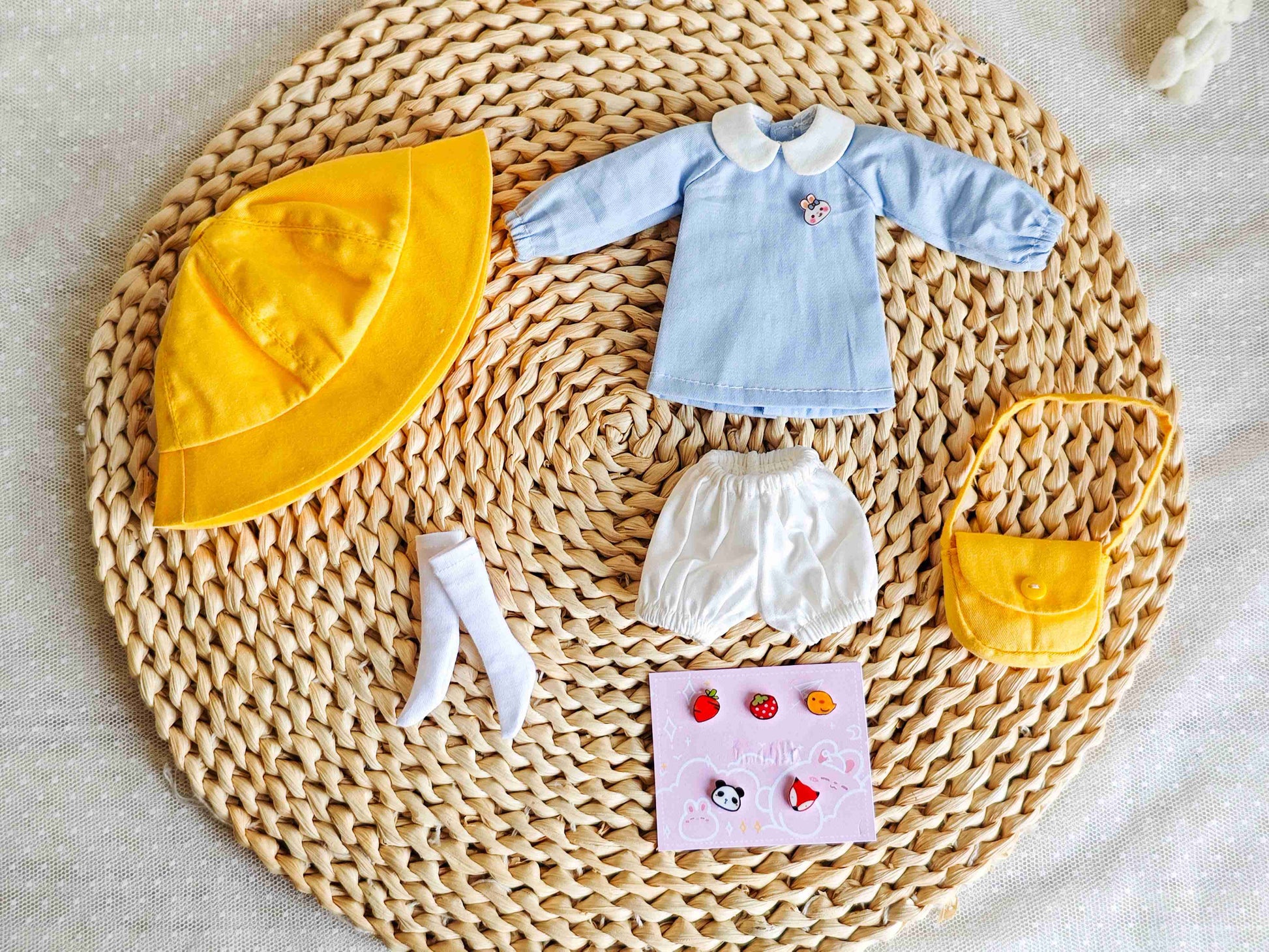 Flat lay product shot of 3-piece doll outfit set including blue smock, yellow hat, and bag.